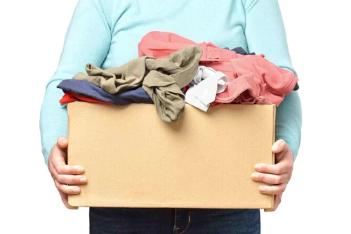 A person in a light turquoise long-sleeved shirt holds a cardboard box of unfolded clothes.