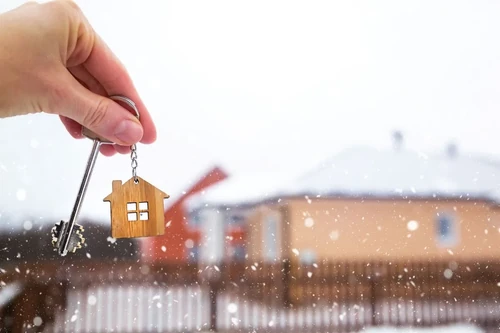 A hand holding a key in front of a house while it snows.