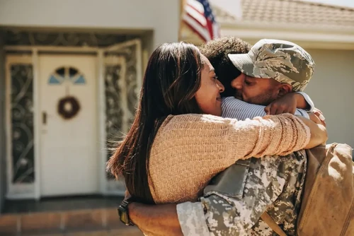 A father in his military uniform hugging his wife and child in front of their house.