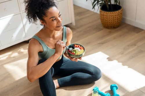 An athletic woman sits on her living room floor, eating a healthy fruit bowl.