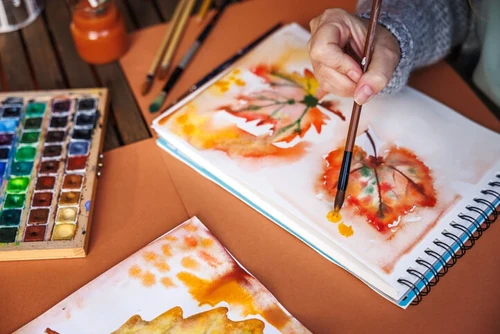 A woman paints a picture of leaves at her desk with supplies for her to use.