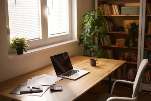 A tidy home office space with a laptop, mug, and business documents sitting on a wooden desk next to a window.