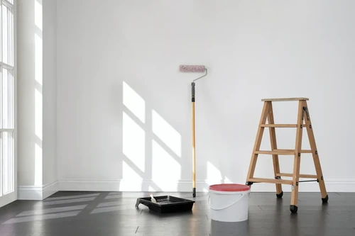 A paint can, plastic pan, brush, and ladder against a white wall in an empty room.